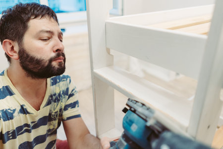 man rubs wooden child bed with power sander or grinder removing old paint
