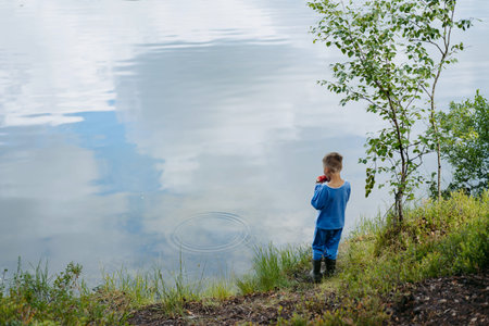 little boy wearing pajamas eating watermelon alone on bank of pure lake. Sky reflecting in waterの写真素材