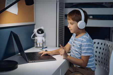 caucasian little boy wearing wireless headphones studying at home using laptop writing in notebook....