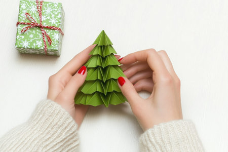 woman hands, wearing white wool sweater, making origami of Christmas tree from green paper on white wooden table, craft gift box near her handsの素材