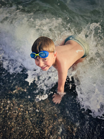 little boy wearing goggles splashing in sea catching waves happily laughingの写真素材