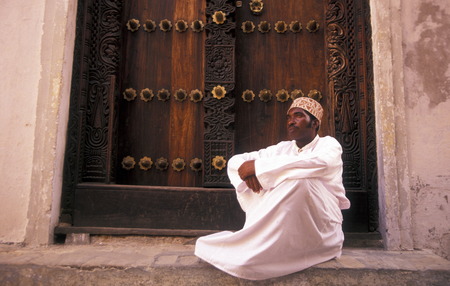 A man from Zanzibar in traditional clothing is heard on an old wooden door in the middle of Stone Town Zanzibar Town capital on the island of Zanzibar which to Tanzaniaのeditorial素材
