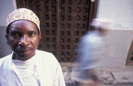 A man from Zanzibar in traditional clothing is heard on an old wooden door in the middle of Stone Town Zanzibar Town capital on the island of Zanzibar which to Tanzaniaのeditorial素材