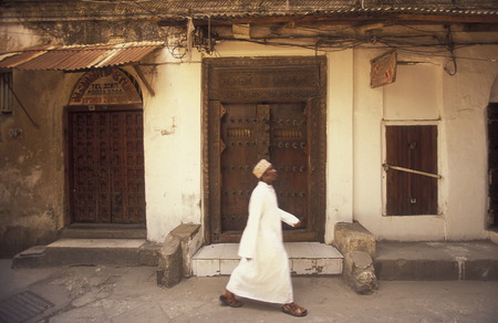 A man from Zanzibar in traditional clothing is heard on an old wooden door in the middle of Stone Town Zanzibar Town capital on the island of Zanzibar which to Tanzaniaのeditorial素材
