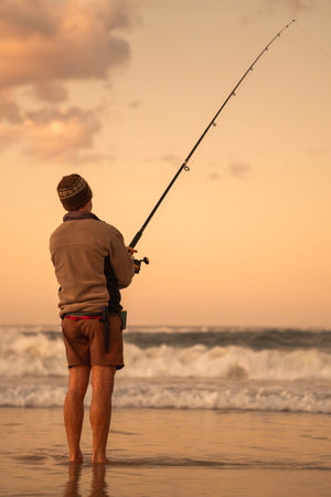 Fisherman with his back fishing on the beach shore during sunriseの写真素材