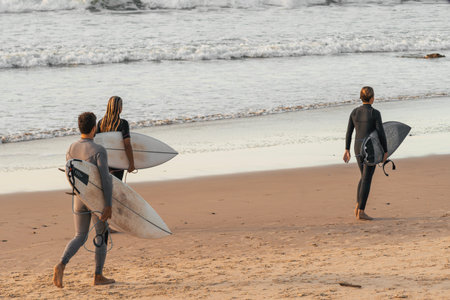 Three surfers walking along the beach with their surfboards dressed in neopreneの写真素材