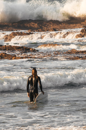 Long-haired surfer in a black wetsuit looking at the aggressive waves of the sea. Verticalの写真素材