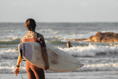 Young surfer in a red wetsuit looking at the sea with the surfboard on his armの写真素材