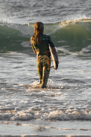 Young surfer girl in camo wetsuit entering the sea holding her surfboard