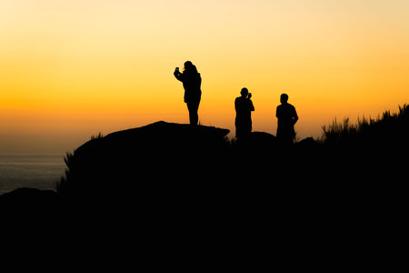 Group of 4 friends watching the sunset on the beach shore and taking photos of the momentの写真素材