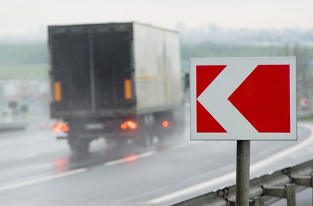 Red and white road alarm sign near sharp turn. Motion-blurred truck on backround.の写真素材