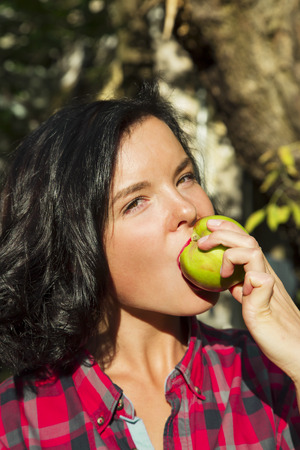 LATVIA, RIGA, SEPTEMBER, 19, 2015 - Portrait of a cute young brunette in a checkered red shirt bites a fresh green apple. Riga, Latviaのeditorial素材