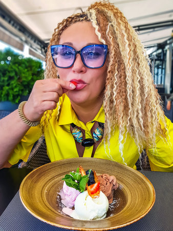 Pretty woman with curly hair in blue glasses eats strawberries from ice cream in Riga, Latvia