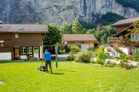SWITZERLAND, WENDEPLATZ, AUGUST 2021 - Active elderly male gardener mow grass with lawn mower against the background of the Swiss Alps, Switzerland.のeditorial素材