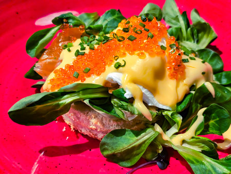 Egg benedict on crispy white bread with Hollandese sauce garnished with red caviar and fresh herbs on a red platter closeupの写真素材