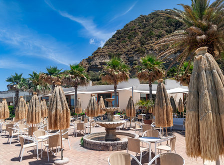Outdoor veranda of a restaurant with tables and straw umbrellas with a fountain at the foot of the mountains on the island of Ischia, Italyの写真素材