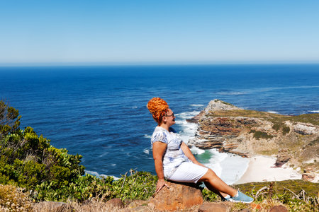 A slender woman with bright red hair sits on the rocky shore of Cape Agulhas, where the Indian and Atlantic Oceans meet, a cape in South Africa, the southernmost point of Africa.の写真素材