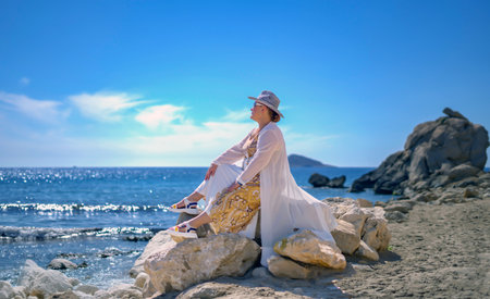 A woman in a long dress with a flowing white cape and a white hat sitting on the rock on the shore of the Mediterranean Sea in Benidorm, Spainの写真素材