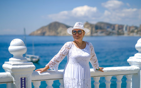 A woman in a long lace dress and a white hat stands near a snow-white staircase and looks at the...