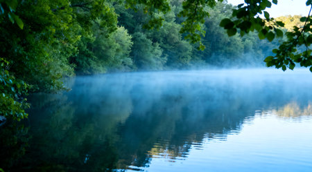 This image depicts a tranquil lake surrounded by dense, vibrant green foliage under the soft glow of morning light. The scene is painted in soothing shades of blue and green, creating a peaceful and artistic representation of nature. Ideal for use in relaxation-themed content, environmental campaigns, or as a calming backdrop in digital media. AI-generated illustration.の素材