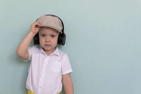 A little boy in a white shirt, yellow shorts and a retro cap listens to music through large headphones.の写真素材