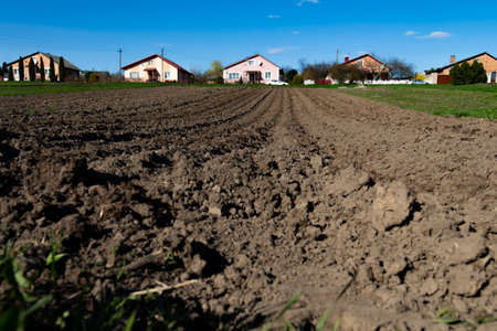 A plowed field for planting crops, potatoes and wheat in early spring on a sunny day. Beds for planting seedlings rise up to a private house and cars.の写真素材