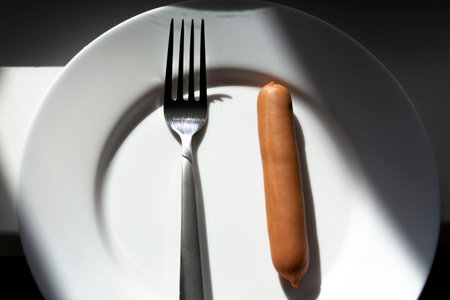 One sausage is impaled on a fork on a white plate with falling sunlight.