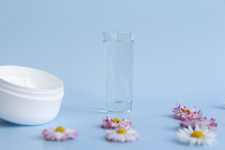 Fresh flowers, daisies are laid out in a corner, with a glass of clean water and a white jar with a cream cosmetic on a blue background and a free space for advertising.の写真素材