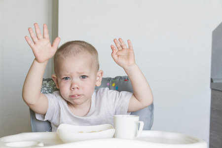 A little boy with a short haircut independently and appetitiously eats food and drinks juice from a tube in a child's chairの写真素材