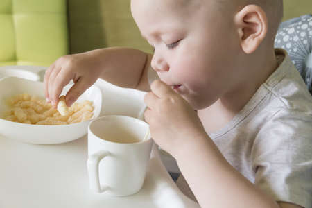 A small child with a short haircut crunches appetitively with corn sticks and drinks juice from a strawの写真素材