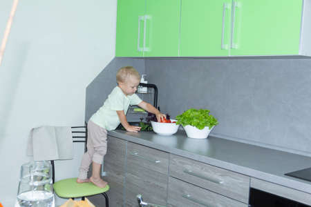 A little boy with a short haircut helps to cook in the kitchen. Washes fresh vegetables and herbs in a black sink in a gray kitchen...の写真素材