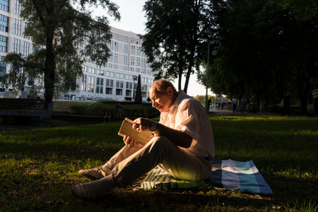 An elderly man in a white shirt is sitting on a blanket, on the ground in a park and reading an...