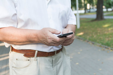 A mobile phone in the large hands of an elderly man in a white shirt on a walk in the park.の写真素材