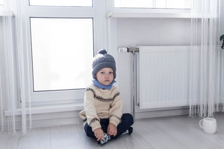 A small child in a sweater and a gray hat is kneeling near a heater with a thermostat...の写真素材