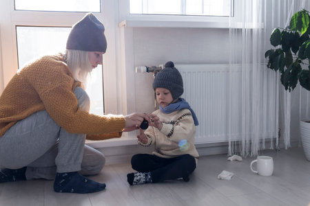 A young mother gives medicine to her little son, who is warmly dressed and sitting on the floor..の写真素材