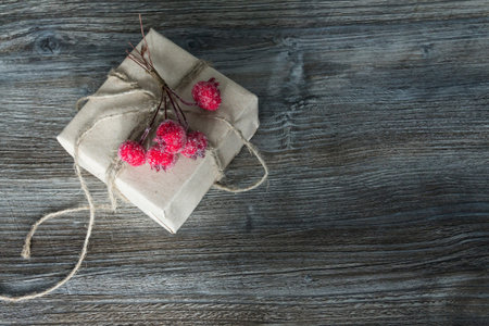 A gift box with red berries, packed in kraft paper and tied with linen twine on a wooden background....