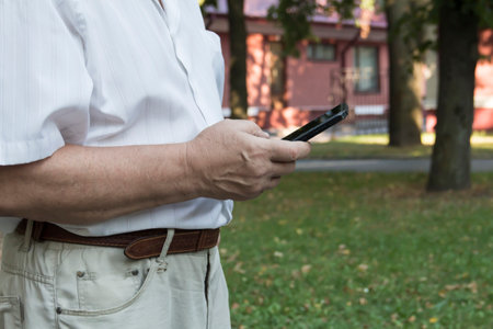 A mobile phone in the large hands of an elderly man in a white shirt on a walk in the park.の写真素材