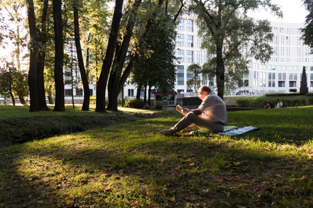 An elderly man in a white shirt is sitting on a blanket, on the ground in a park and reading an interesting book. A pensioner alone is resting in nature, passionate about his hobbyの写真素材