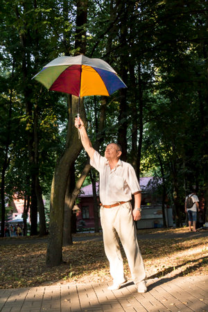 An elderly man stands under a large, multicolored umbrella. A pensioner on a walk in the park.の写真素材