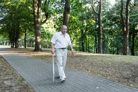 An old, stooped man walks sullenly and alone along the path in the park with a gray umbrella instead of a cane..の写真素材