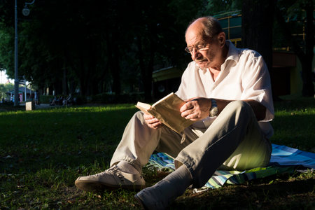 An elderly man in a white shirt is sitting on a blanket, on the ground in a park and reading an interesting book. A pensioner alone is resting in nature, passionate about his hobby.の写真素材