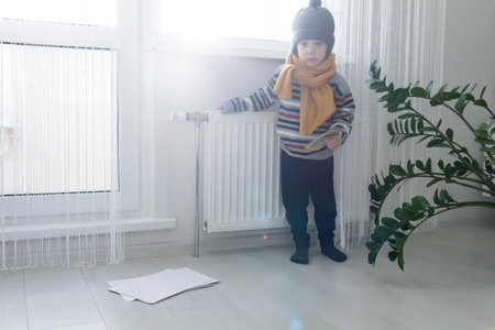 The child is warmly dressed in a sweater and a hat, standing and holding on to the radiator with money and bills. The concept of the economic crisis and the lack of heat and heating in homesの写真素材