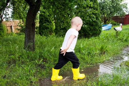 A small, bald-headed boy in yellow boots runs in the countryside through puddles in the fresh airの写真素材