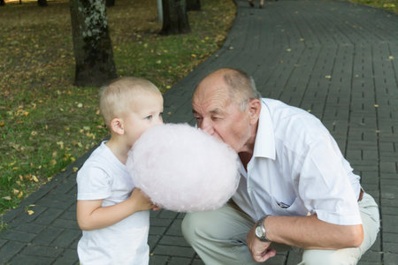 An elderly man with his mouth wide open greedily bites cotton candy. The grandson is eating cotton candy next to his grandfather.の写真素材