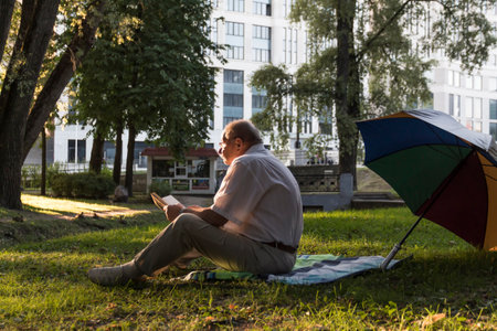 An elderly man in a white shirt is sitting on a blanket, on the ground in a park and reading an interesting book. A pensioner alone is resting in nature, passionate about his hobbyの写真素材