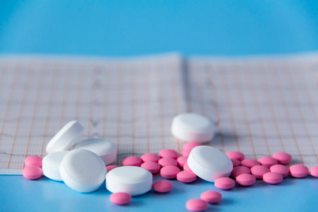 A large handful of pink and white medicines lie on the electrocardiogram of the heart, on a blue background. The concept of a healthy lifestyle and timely medical examinationの写真素材