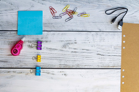 Blue sheets for notes, colored paper clips, a sheet of craft paper, paper clips, a charger for gadgets and a corrector for office work and school on a light, wooden background.の写真素材