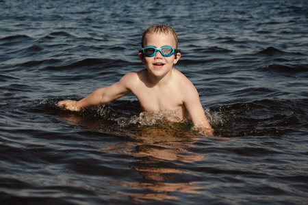 child happily excitedly enters water, swims, dives lake or pond on a hot, sunny summer day during vacation or school break. active healthy lifestyle. child wearing turquoise swimming trunks goggles.の写真素材