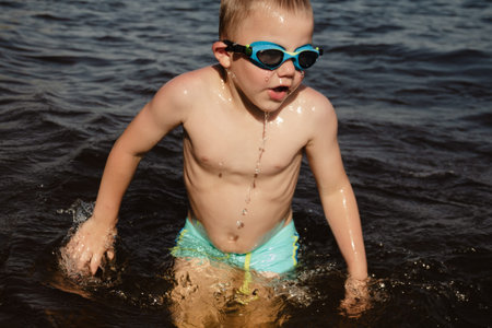 child happily excitedly enters water, swims, dives lake or pond on a hot, sunny summer day during vacation or school break. active healthy lifestyle. child wearing turquoise swimming trunks goggles.の写真素材