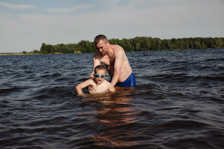father and son swimming in a lake. man stands calmly in the water nearby. In the background is a green shore, calm waters, and a clear sky. The atmosphere evokes summer relaxation and family warmth.の写真素材